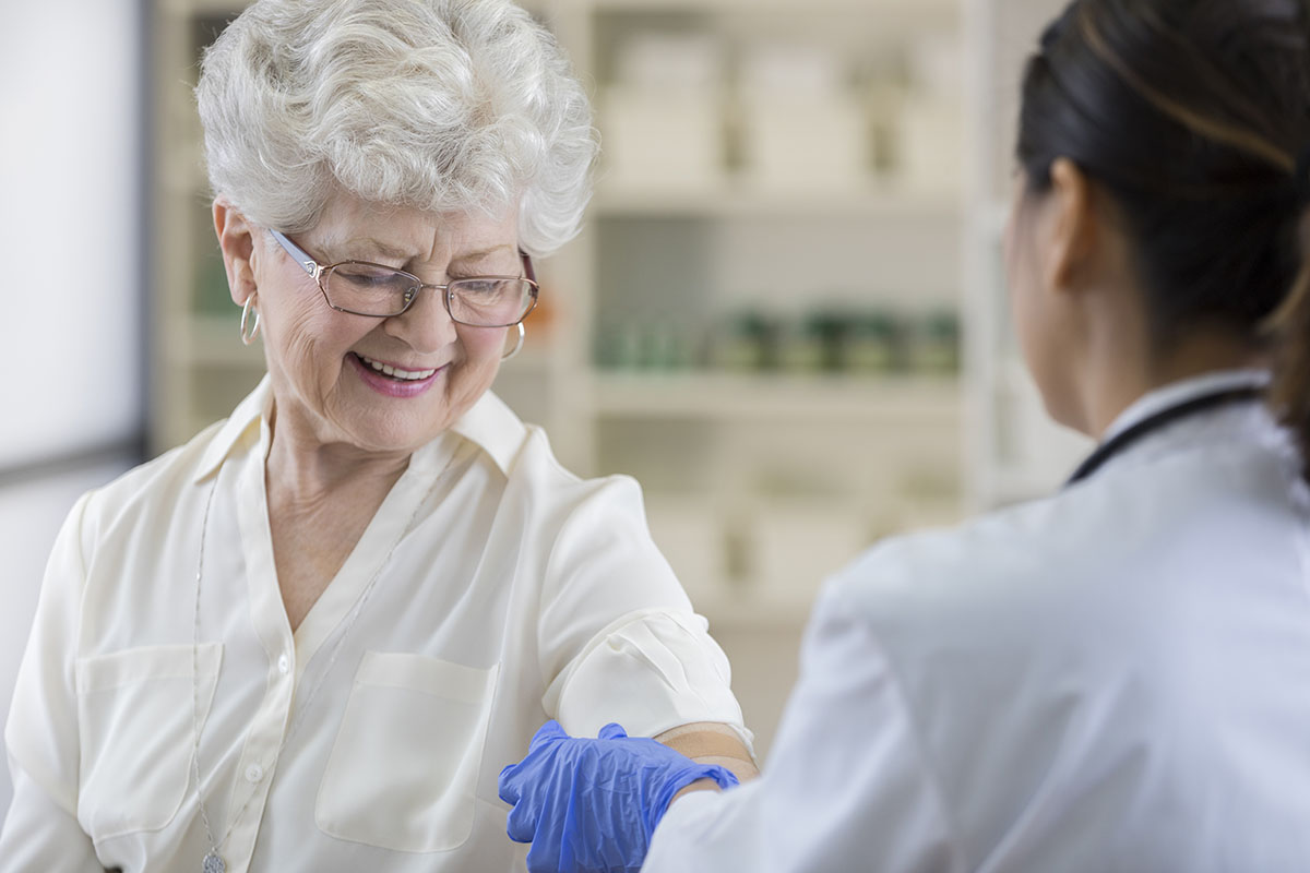 A smiling senior pharmacy customer looks down at her upper arm as a gloved pharmacist places a band-aid over her flu shot site.  There are shelves in the background.