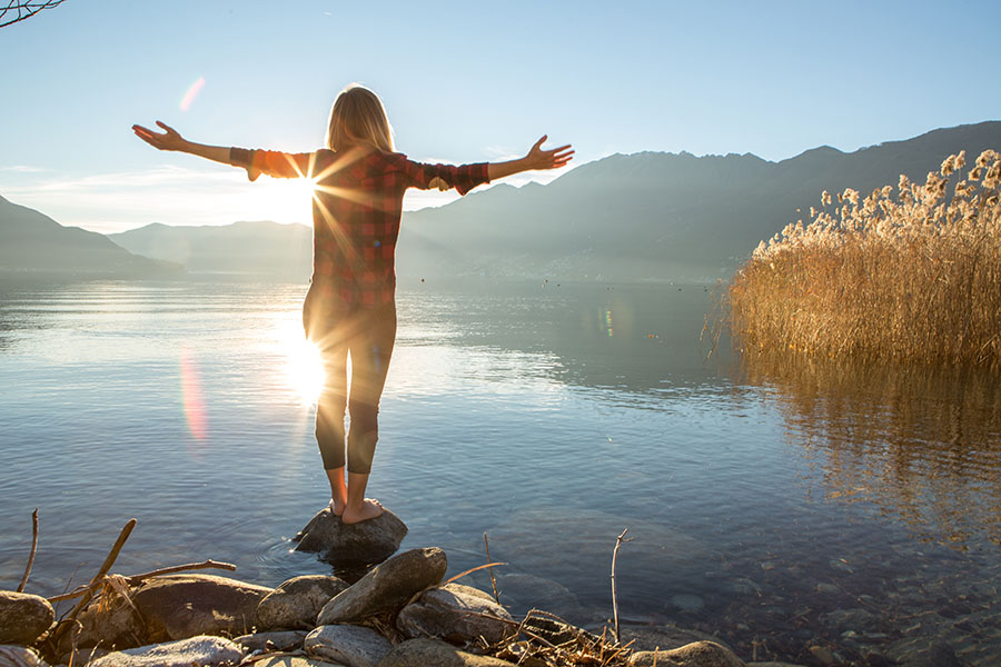 Young cheerful woman by the lake enjoying nature. Arms outstretched for positive emotion.